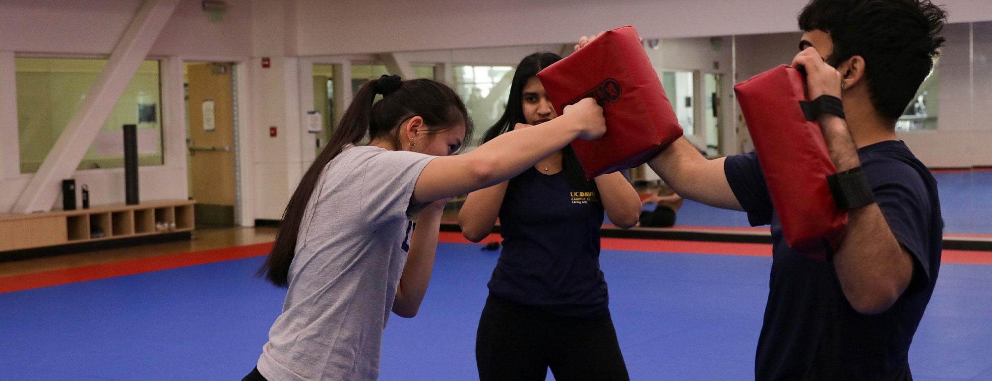 A trainer shows a student how to punch a pad, which a third student holds up for practice. 