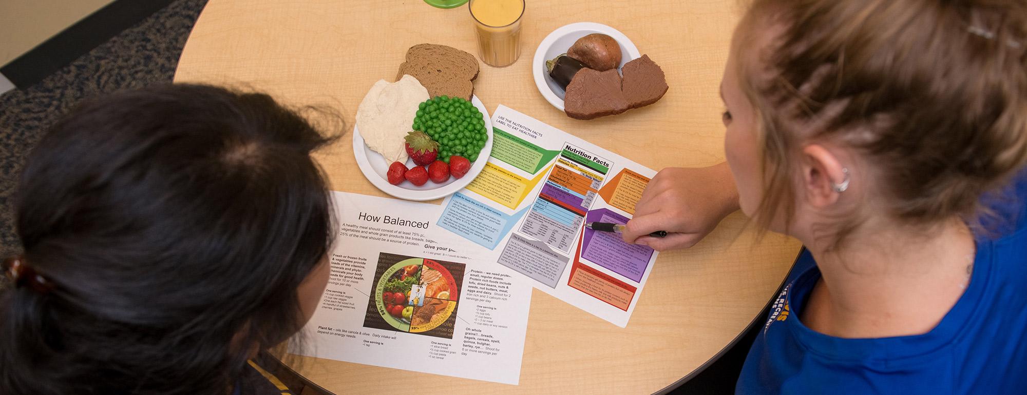 Two students look at a nutrition plan on a table with a sample meal on it